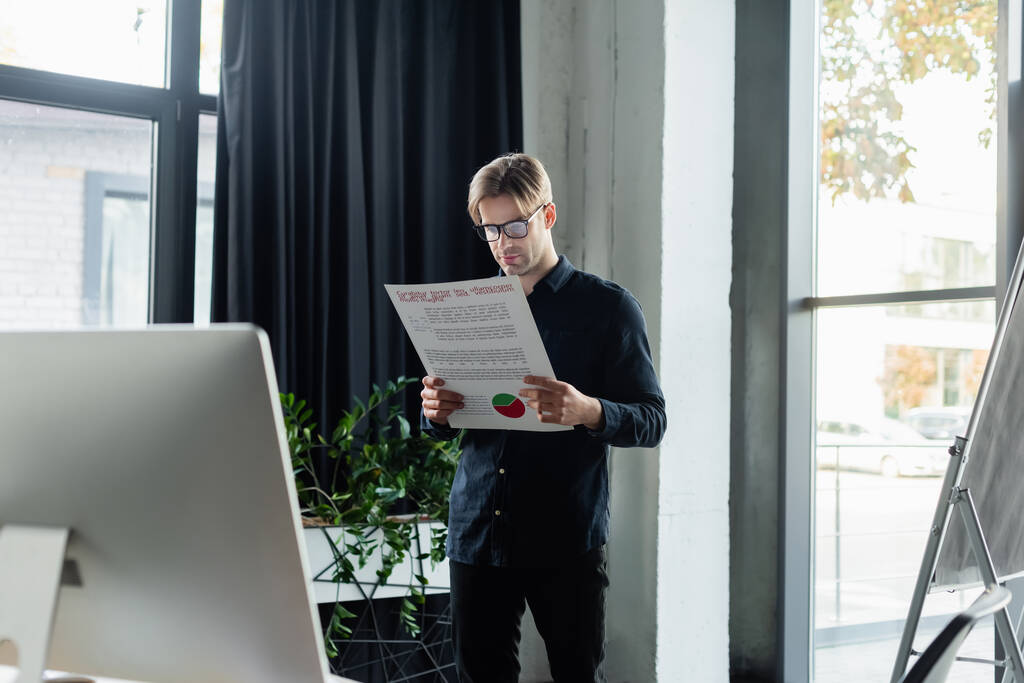 Програміст в окулярах тримає папір біля комп'ютера і фліп-чарт в офісі
  - Фото, зображення