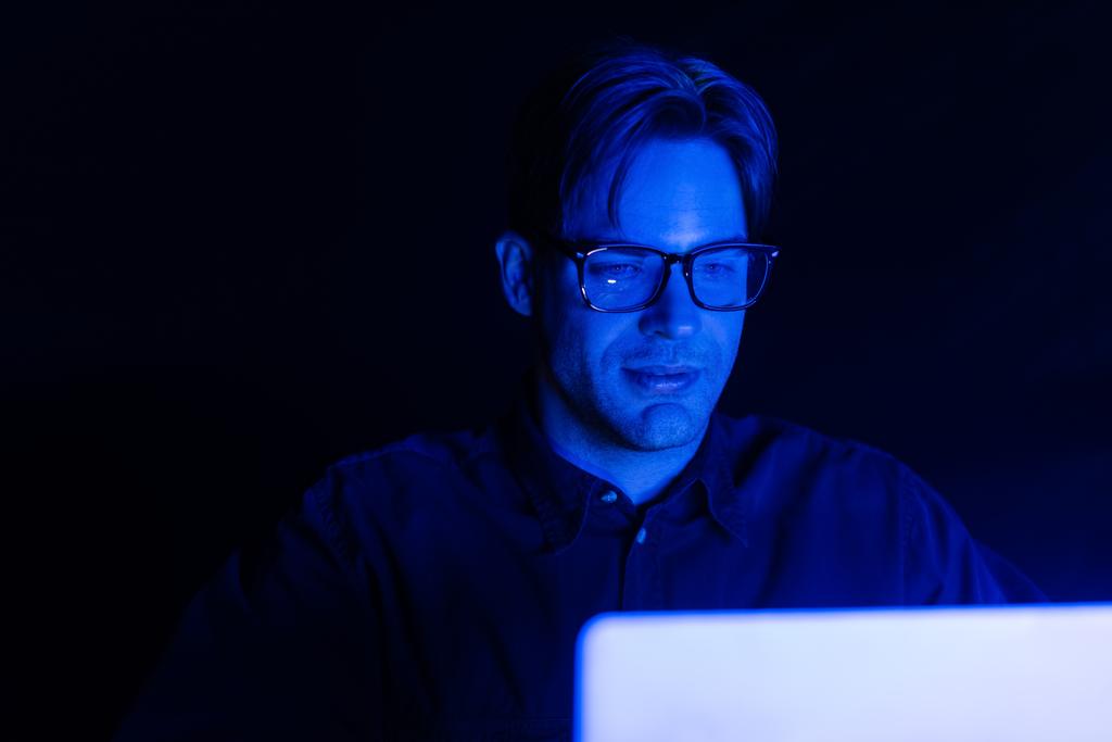 Programista w okularach patrząc na zamazany laptop odizolowany na czarno  - Zdjęcie, obraz