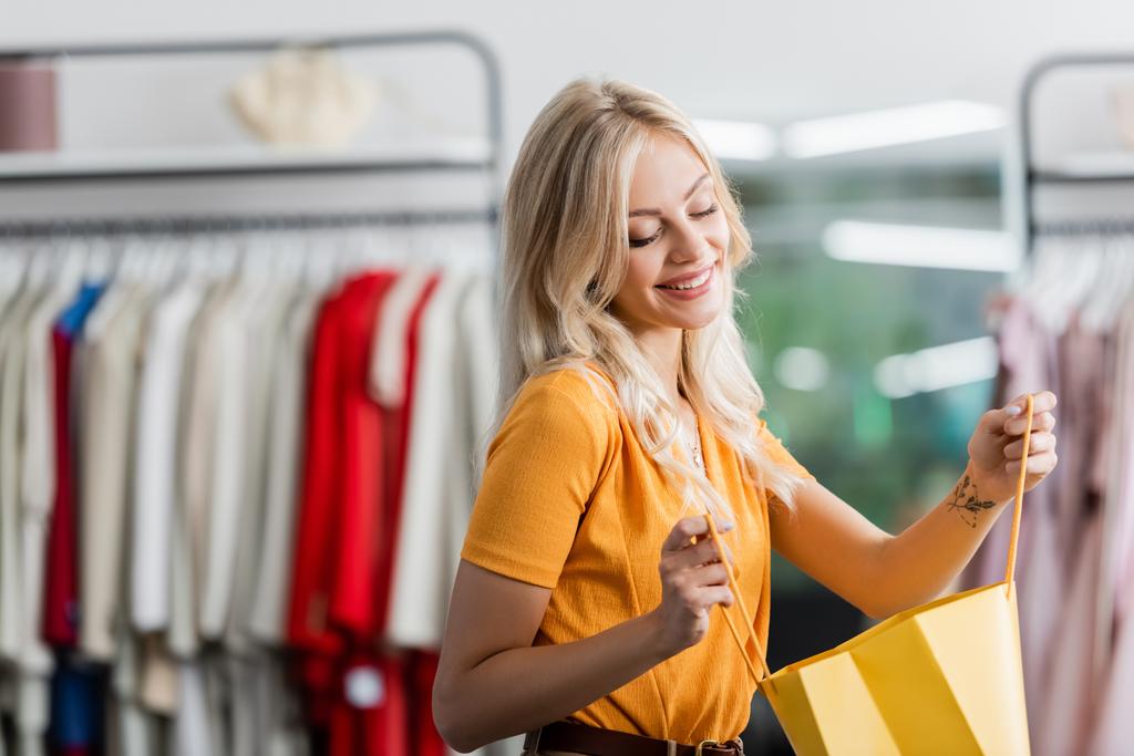 happy woman looking at yellow shopping bag in clothing boutique - Photo, Image