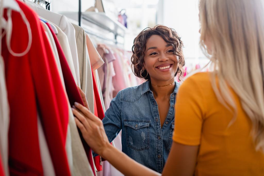 onnellinen afrikkalainen amerikkalainen myymälä avustaja katselee blondi asiakas boutique  - Valokuva, kuva
