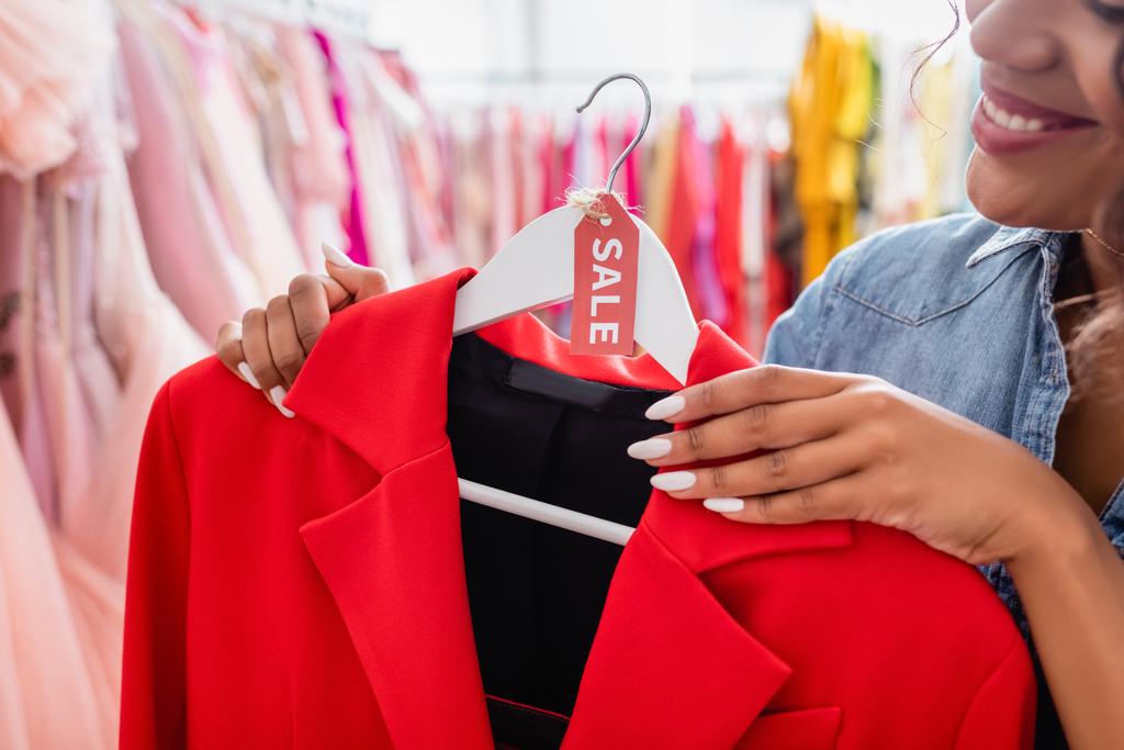 vista ritagliata di felice assistente negozio africano americano con giacca rossa in vendita tag in boutique - Foto, immagini