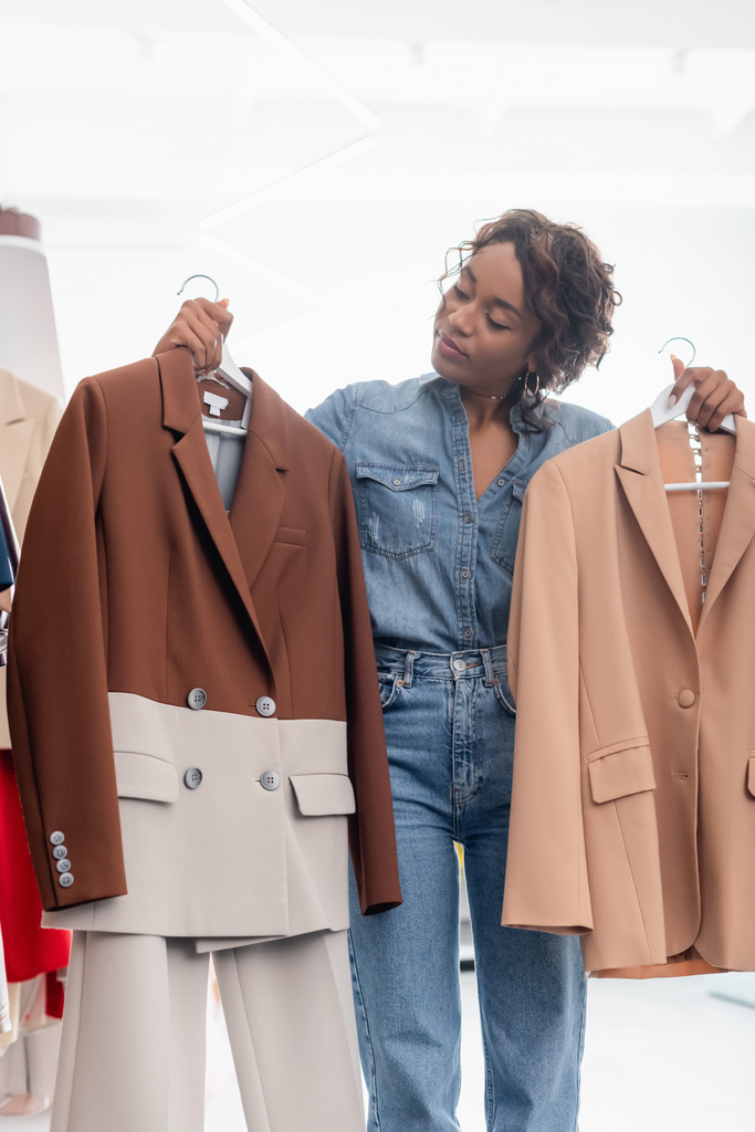 mujer afroamericana rizada sosteniendo perchas con chaquetas en boutique - Foto, Imagen