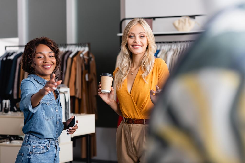 femme heureuse tenant tasse en papier tandis que l'assistant de magasin afro-américain avec smartphone pointant loin  - Photo, image