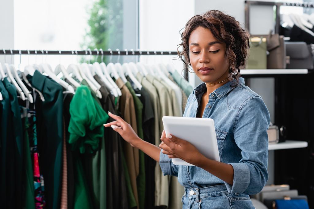 молодая африканская американская продавщица, держащая цифровой планшет и указывая на одежду на стойке - Фото, изображение