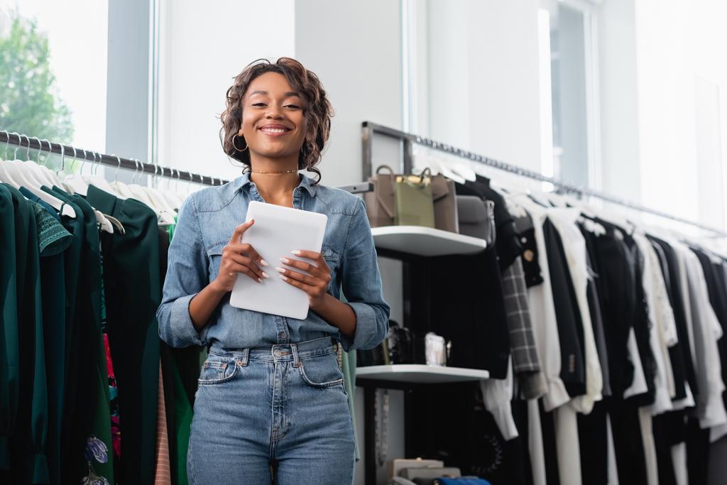 vendedora afroamericana positiva sosteniendo tableta digital cerca de la ropa en rack - Foto, imagen