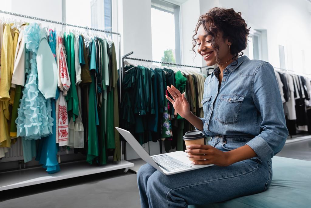 happy african american woman waving hand during video chat in clothing boutique - Photo, Image