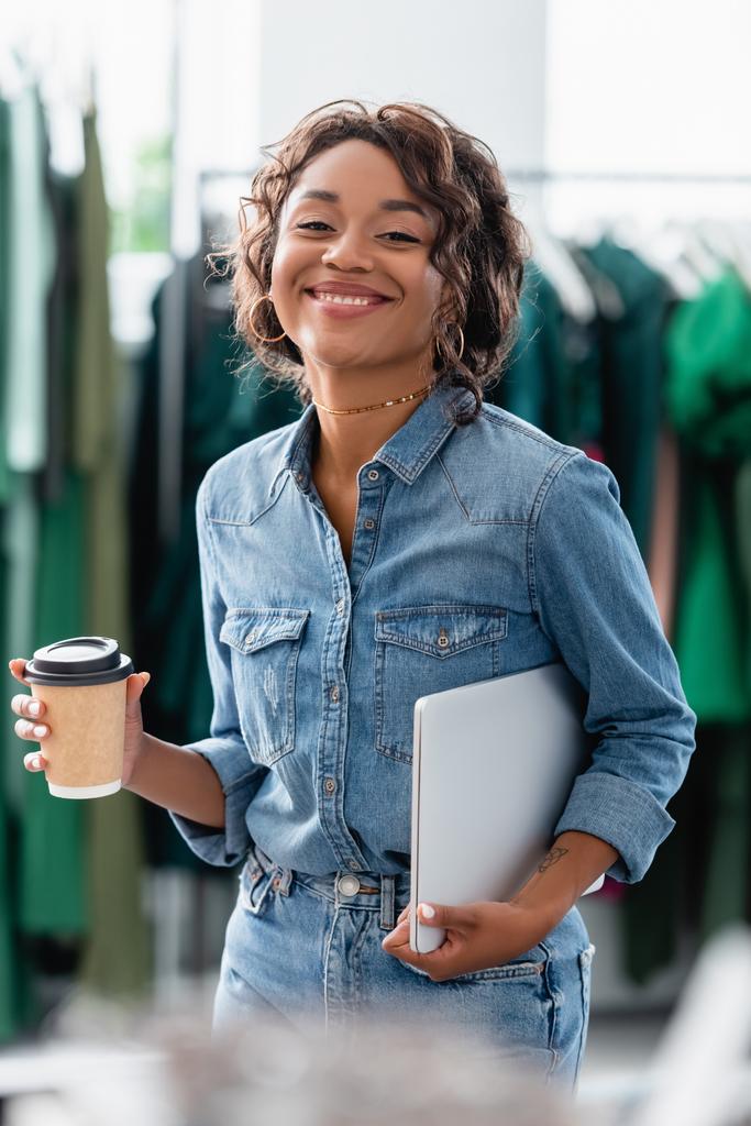 alegre mujer afroamericana sosteniendo portátil y taza de papel en boutique  - Foto, Imagen