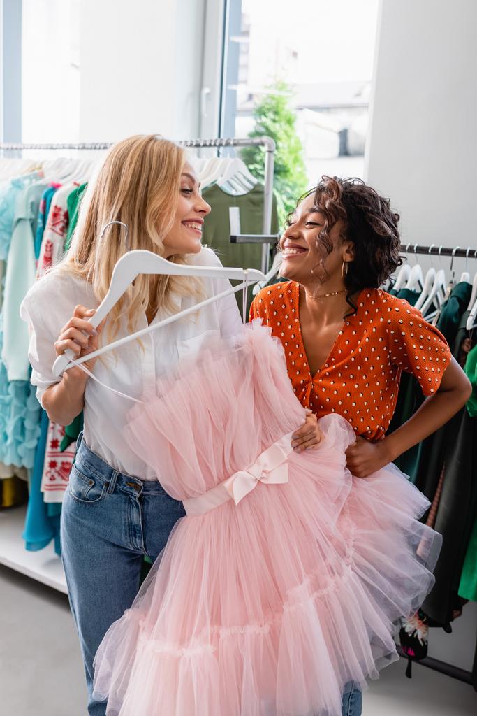 happy blonde woman holding pink dress and looking at joyful african american friend  - Photo, Image