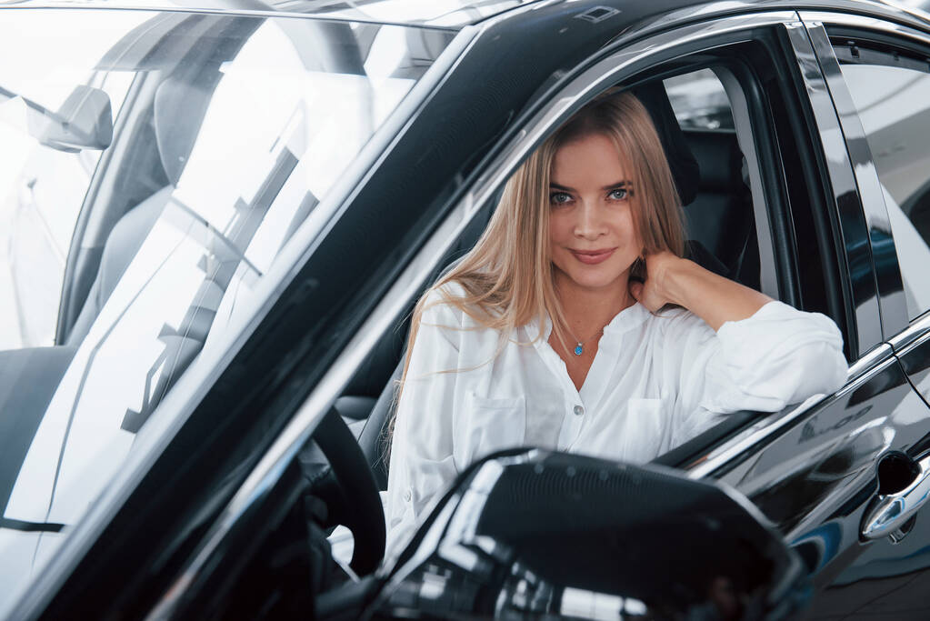 In een wit shirt. Mooi blond meisje zitten in de nieuwe auto met moderne zwarte interieur. - Foto, afbeelding