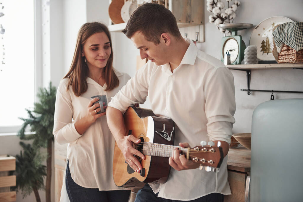 Ρομαντικό πρωινό στο σπίτι. Νεαρός κιθαρίστας παίζει ερωτικό τραγούδι για την κοπέλα του στην κουζίνα. - Φωτογραφία, εικόνα