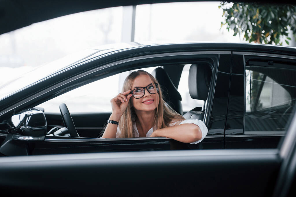 Vihreä kasvi takana. Kaunis blondi tyttö istuu uudessa autossa, jossa on moderni musta sisustus. - Valokuva, kuva