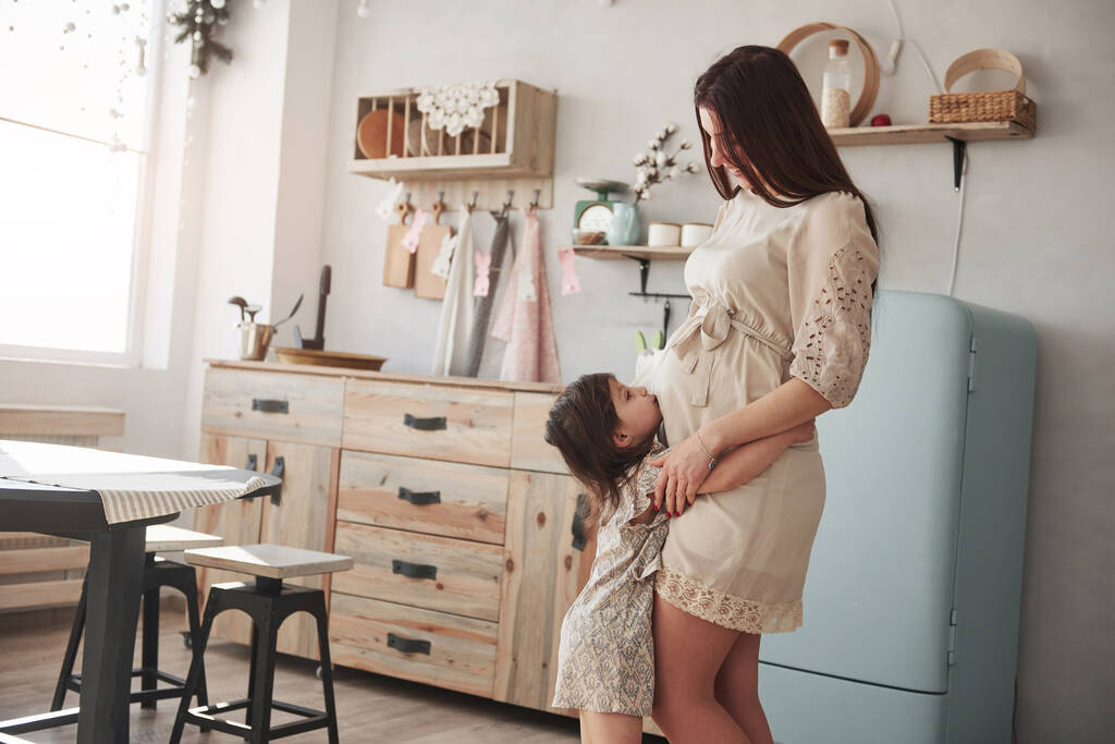 Ребенок целует живот. Молодая положительная брюнетка стоит на кухне днем. - Фото, изображение