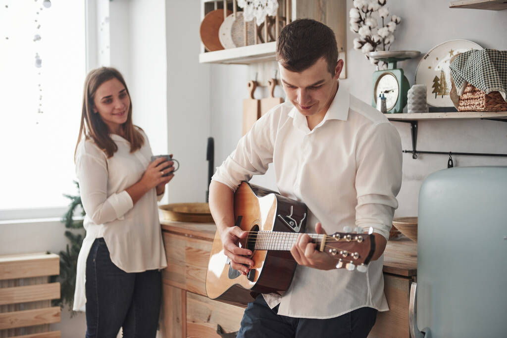 Jonge gitarist speelt liefdeslied voor zijn vriendin in de keuken. - Foto, afbeelding