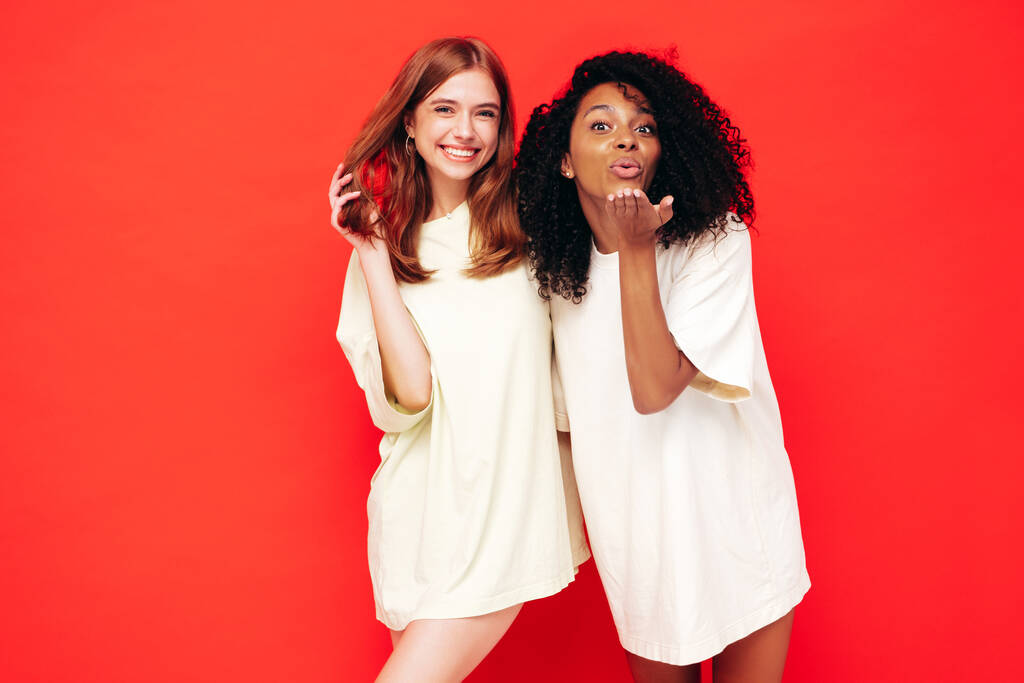 Due giovani belle donne hipster internazionali sorridenti in abiti estivi alla moda. Sexy donne spensierate posa vicino muro rosso in studio. Modelli positivi divertirsi. Concetto di amicizia - Foto, immagini