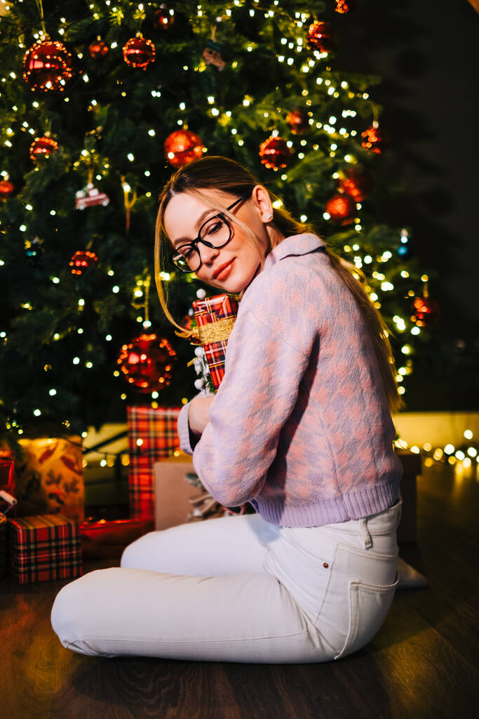 Happy caucasian woman holding big Christmas present near Christmas tree. - Photo, Image