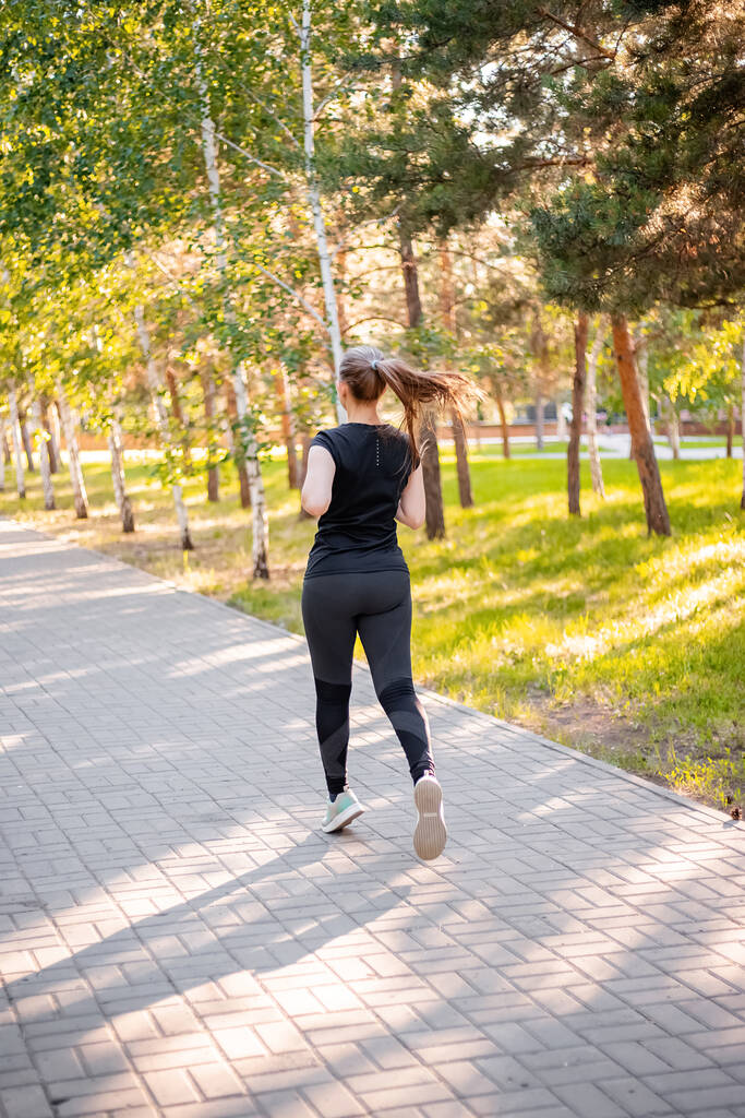 Ein Mädchen in schwarzer Sportuniform joggt im Abendpark. Selbstsportkonzept.  - Foto, Bild