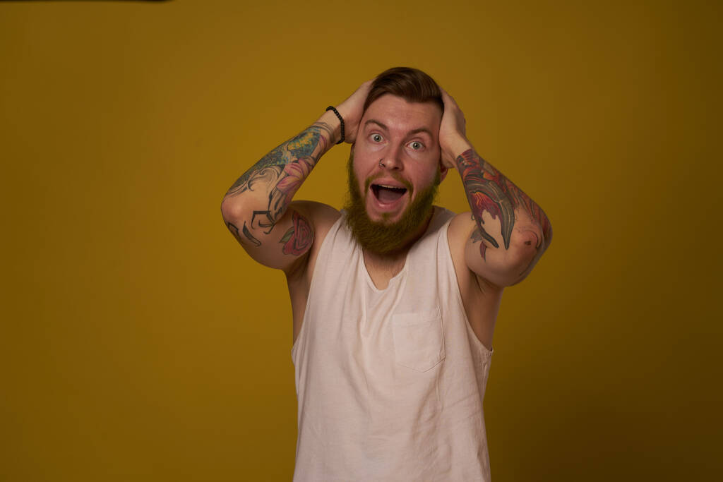 γενειοφόρος άντρας με λευκό μπλουζάκι με τατουάζ στα χέρια του, με σοβαρή εμφάνιση. - Φωτογραφία, εικόνα