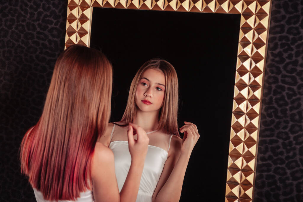 Portrét docela roztomilé dospívající dívky v elegantních šatech v zrcadle v tmavém stylovém interiéru obývacího pokoje. Dětské emoce a pózování. Koncept stylu, módy a krásy. Kopírovat místo pro web - Fotografie, Obrázek
