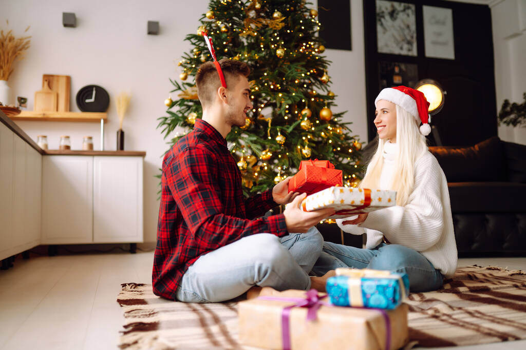 Noel hediyesi değişimi. Genç çift Noel ağacının yakınındaki rahat oturma odasında hediye kutusu açıyor. Romantik bir gün. Kış tatili. - Fotoğraf, Görsel