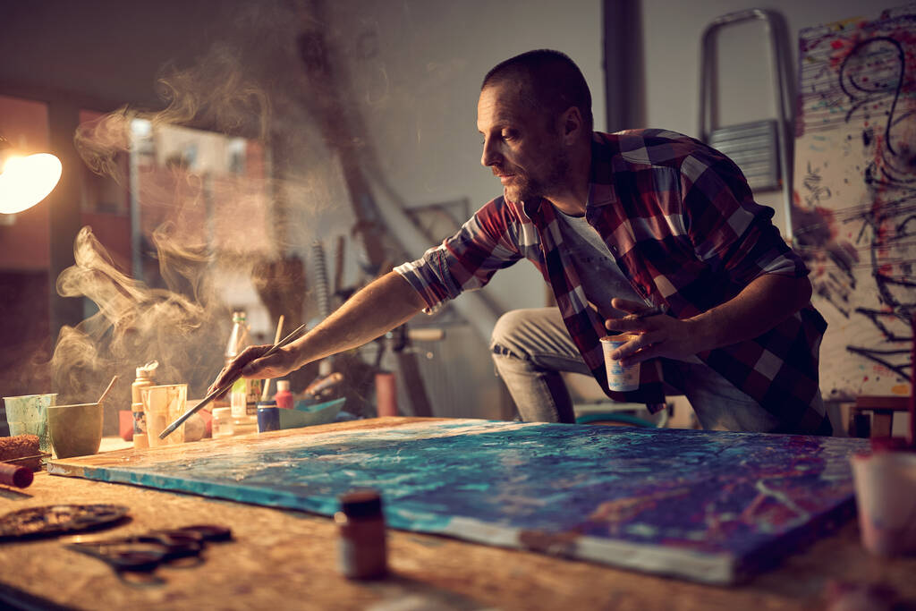 Ένας νεαρός άνδρας καλλιτέχνης σε μια δημιουργική ατμόσφαιρα στο στούντιο επικεντρώνεται σε ένα έργο για τη νέα ζωγραφική του. Τέχνη, ζωγραφική, στούντιο - Φωτογραφία, εικόνα