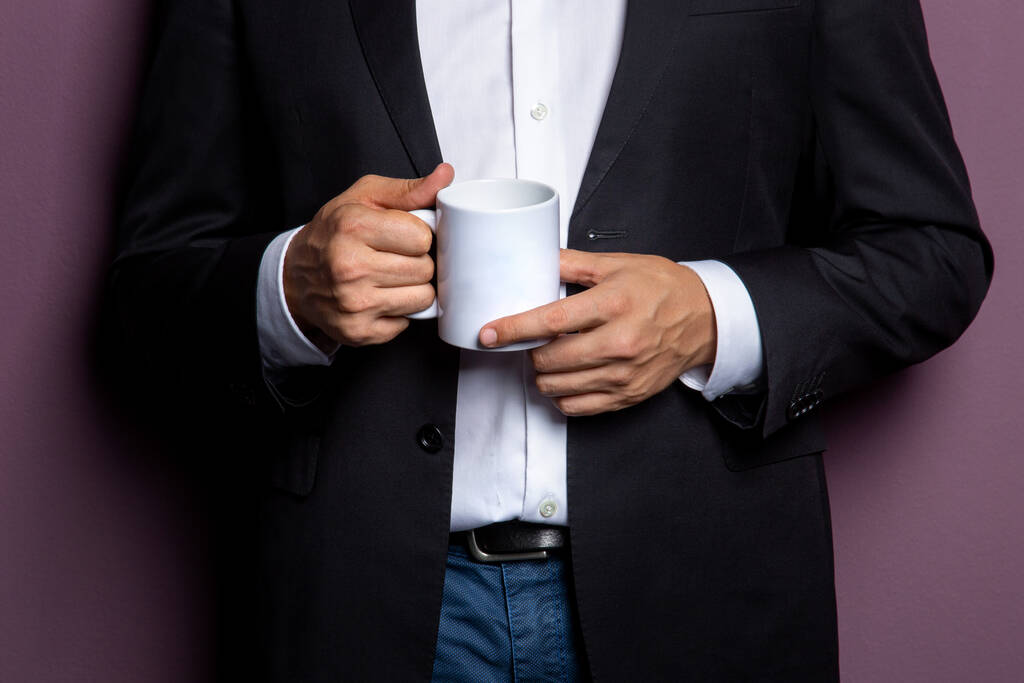 een man in een zwart jasje houdt een witte mok thee in zijn handen. Vrije ruimte op de mok voor de inscriptie. concept is een voorbeeld voor een bedrijfslogo. - Foto, afbeelding