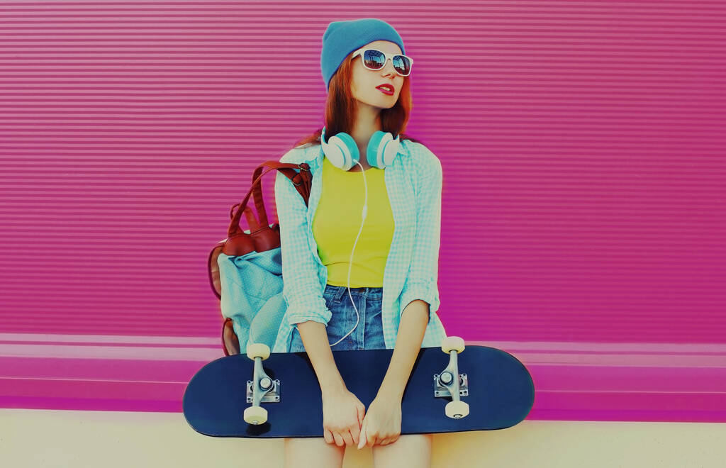 Summer portrait of young woman with skateboard listening to music in headphones on colorful pink background - Photo, Image