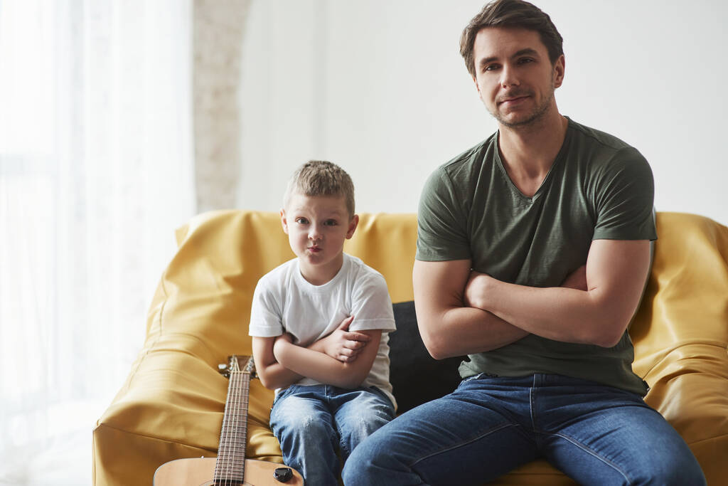 Κάνει αστεία γκριμάτσα. Ο πατέρας και ο γιος του κάθονται στον καναπέ μαζί. Ακουστική κιθάρα στο κρεβάτι. - Φωτογραφία, εικόνα