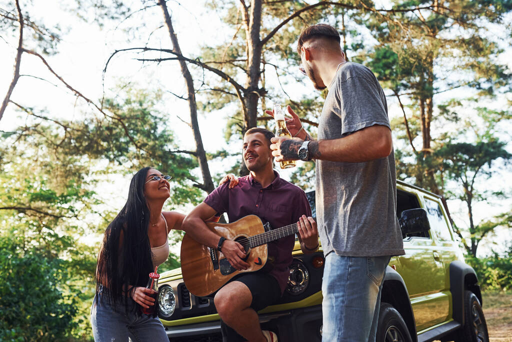 Cantando canciones juntos. Amigos tienen buen fin de semana al aire libre cerca de su coche verde con guitarra acústica. - Foto, imagen