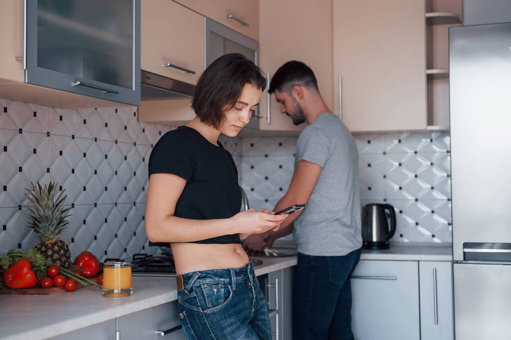 Lire quelques nouvelles sur smartphone. Jeune couple dans la cuisine moderne à la maison à leur week-end le matin. - Photo, image