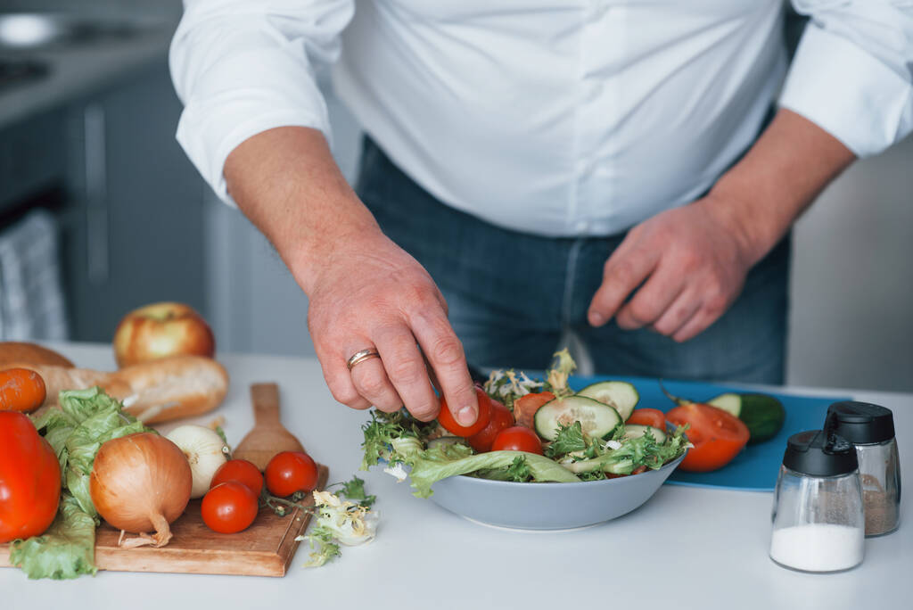 Kis paradicsomot teszek a salátába. Férfi fehér ingben ételt készít a konyhában zöldségekkel. - Fotó, kép