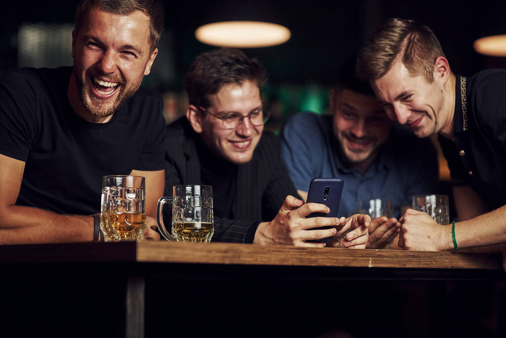 Contenido divertido en smartphone. Tres amigos descansando en el pub con cerveza en las manos. Tener conversación. - Foto, imagen