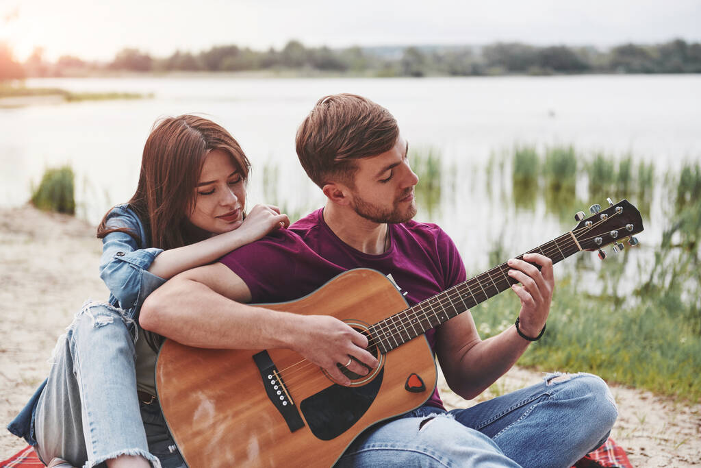 Muž hraje na kytaru pro svou přítelkyni na pláži na jejich piknik ve dne. - Fotografie, Obrázek