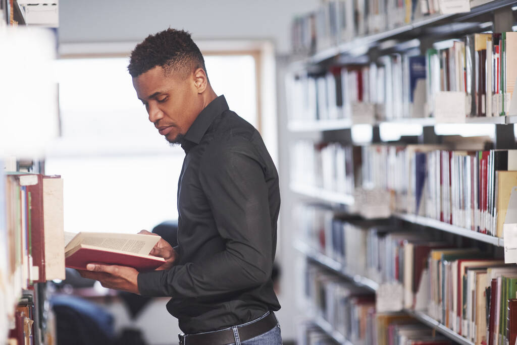 Dobre naturalne oświetlenie. Afrykański Amerykanin w bibliotece szuka informacji w książkach.. - Zdjęcie, obraz