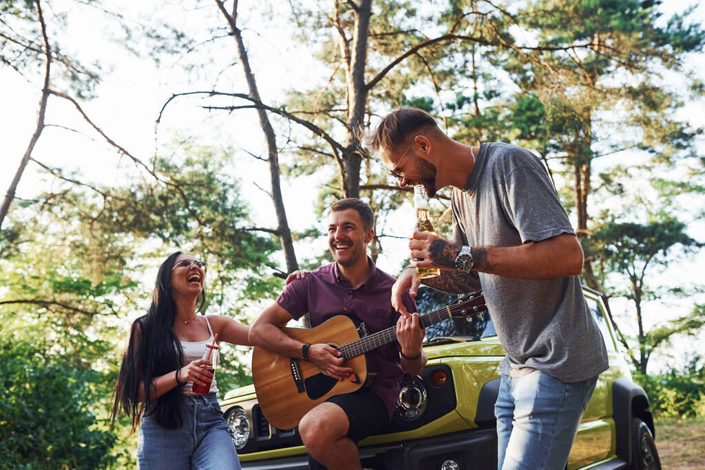 Cantando canciones juntos. Amigos tienen buen fin de semana al aire libre cerca de su coche verde con guitarra acústica. - Foto, Imagen