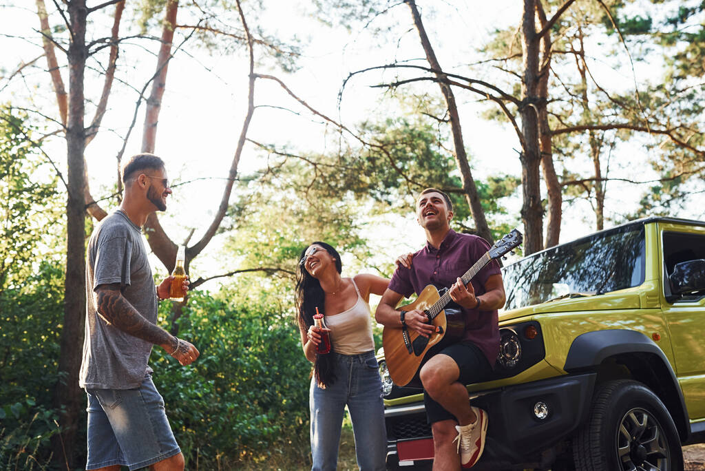 Cantando canzoni insieme. Gli amici hanno un bel fine settimana all'aperto vicino alla loro auto verde con chitarra acustica. - Foto, immagini