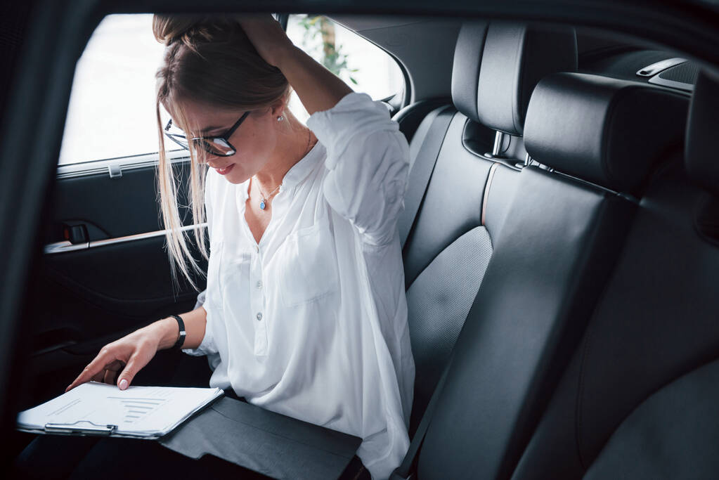 Notizblock liegt auf den Beinen. Kluge Geschäftsfrau sitzt auf dem Rücksitz des Luxusautos mit schwarzem Interieur. - Foto, Bild