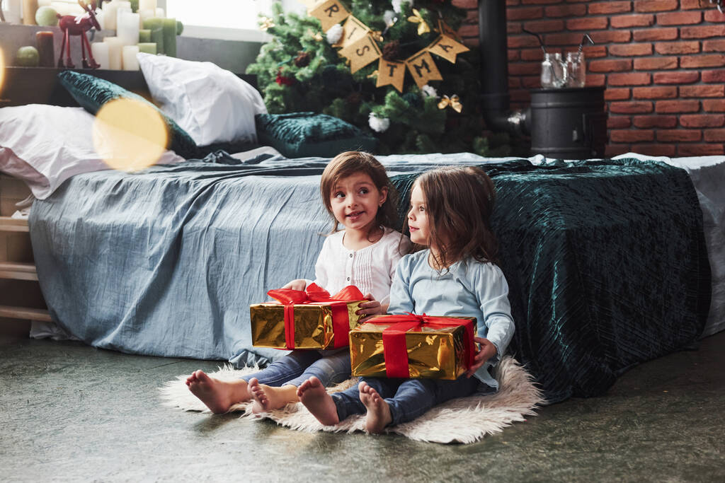 Upřímné emoce. Vánoční prázdniny s dárky pro tyto dvě děti, které sedí uvnitř v pěkném pokoji u postele. - Fotografie, Obrázek