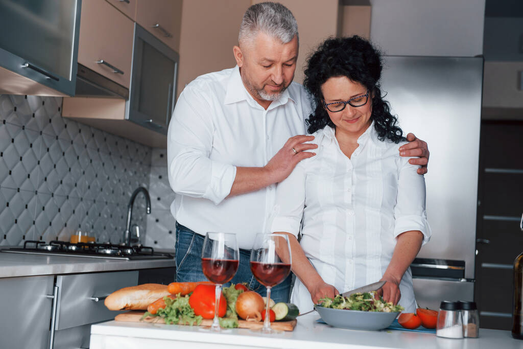 Happy together. Man and his wife in white shirt preparing food on the kitchen using vegetables. - Photo, Image