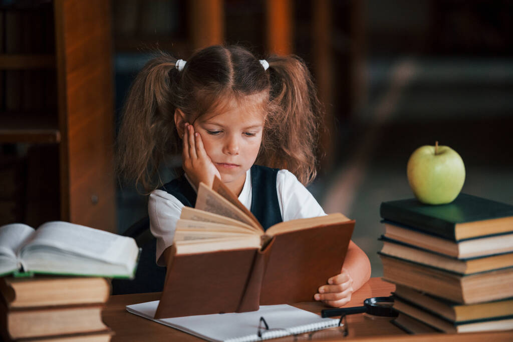 Устал и устал. Милая маленькая девочка с косичками в библиотеке. Apple на книгах. - Фото, изображение
