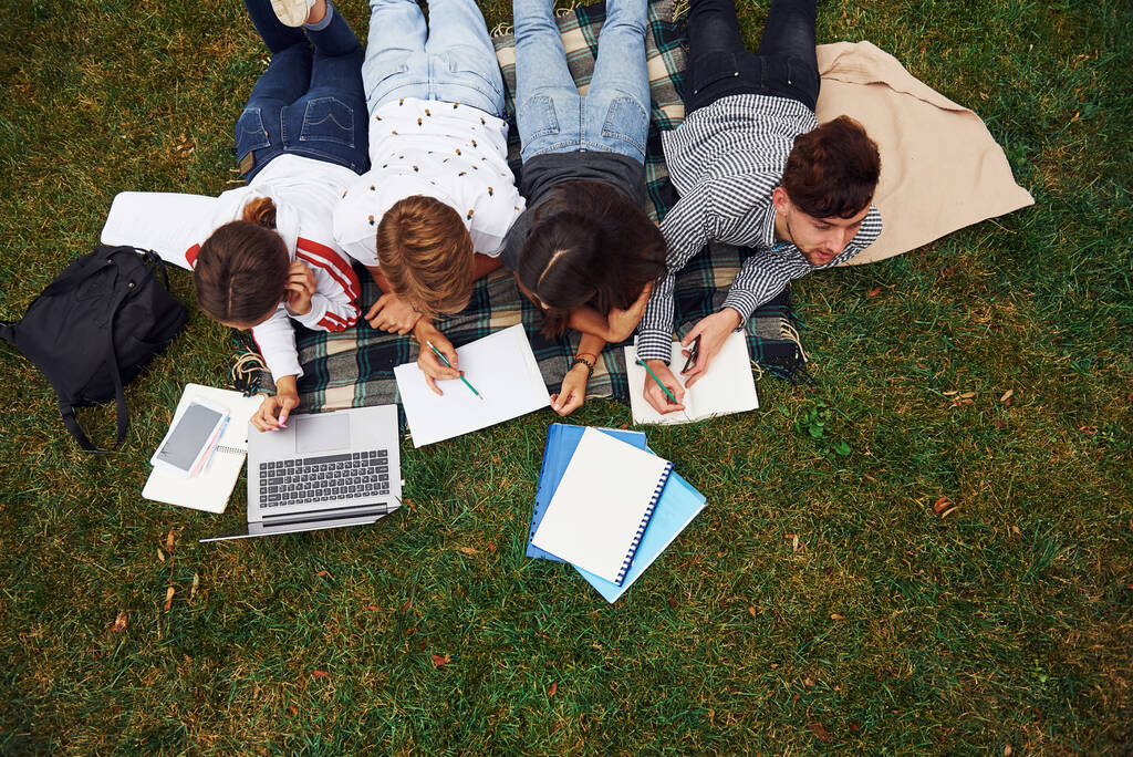 J'écris sur papier. Avec ordinateur portable. Groupe de jeunes étudiants en tenue décontractée sur herbe verte le jour. - Photo, image