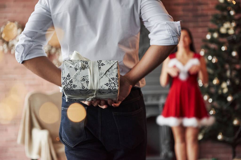 Close up view. Man stands and holds gift box behind. Woman in red dress will now receive Christmas gift from boyfriend. - Photo, Image