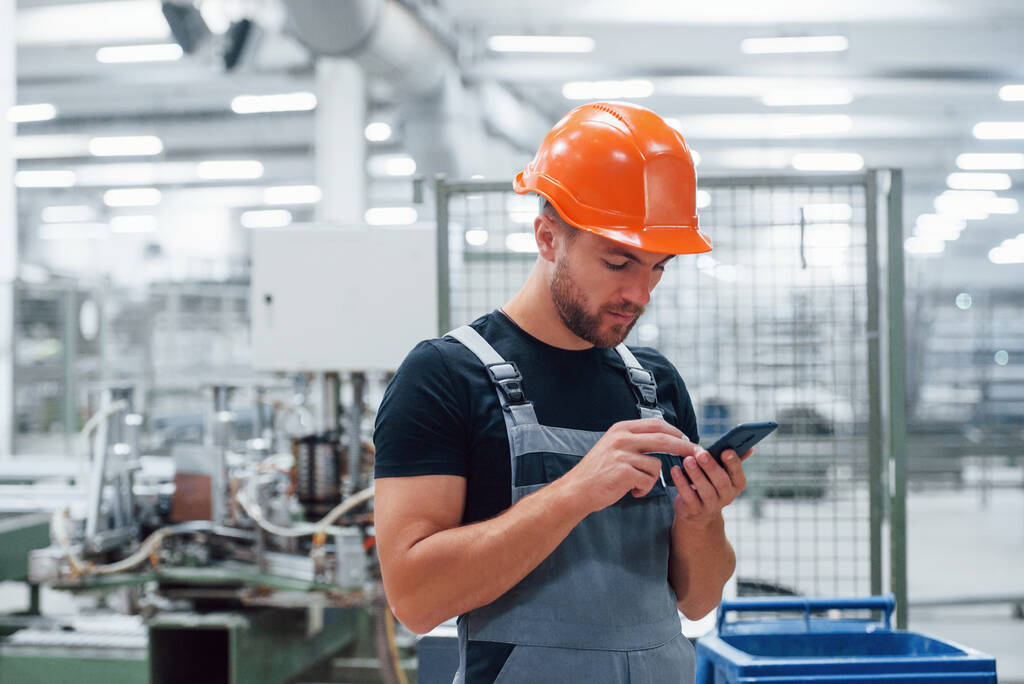 Smartphone in handen. Industriële werknemer binnen in de fabriek. Jonge technicus met oranje harde hoed. - Foto, afbeelding