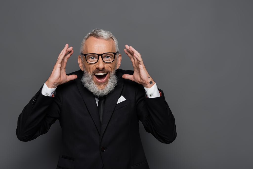 изумленный бизнесмен средних лет в очках жестикулирует на сером - Фото, изображение