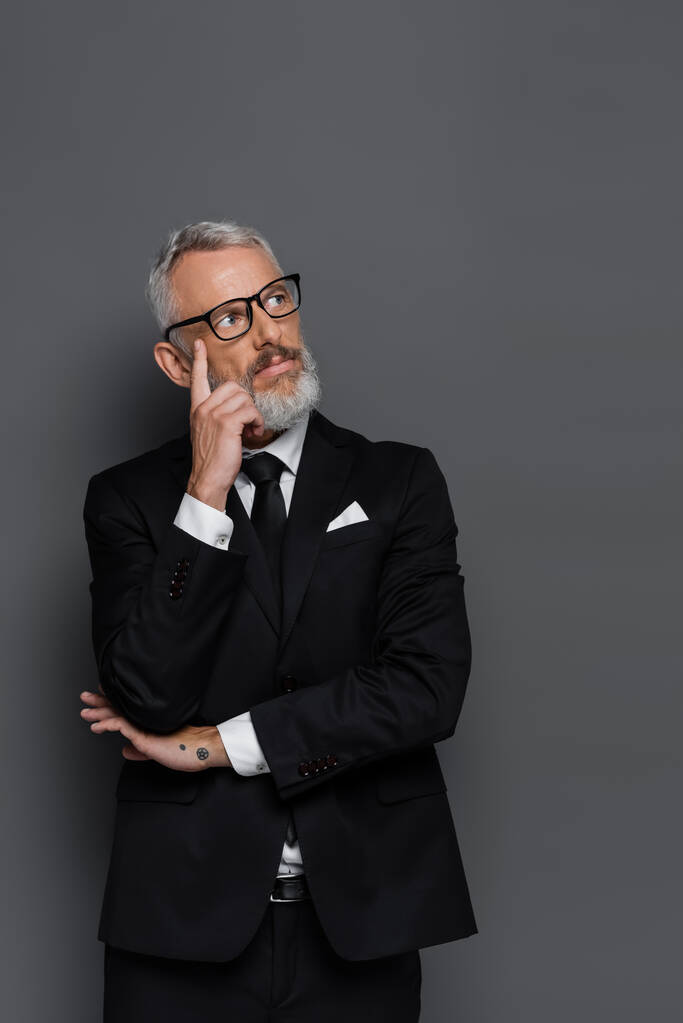 задумчивый бизнесмен средних лет в очках, смотрящий в сторону, изолированный от серого - Фото, изображение