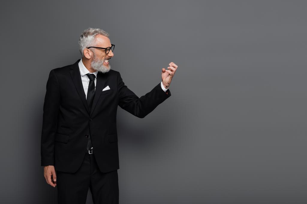 πλαϊνή άποψη του θυμωμένος μεσήλικας επιχειρηματίας σε γυαλιά gesturing απομονώνονται σε γκρι - Φωτογραφία, εικόνα