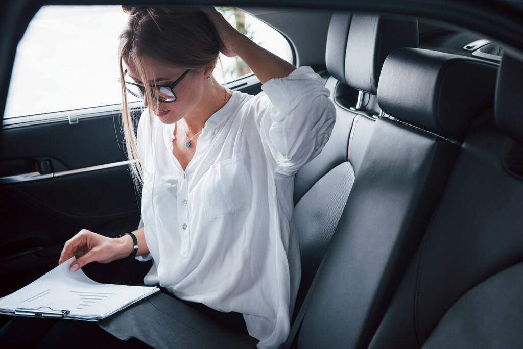 Ważne dokumenty. Smart businesswoman siedzi na tylnym siedzeniu luksusowego samochodu z czarnym wnętrzem. - Zdjęcie, obraz