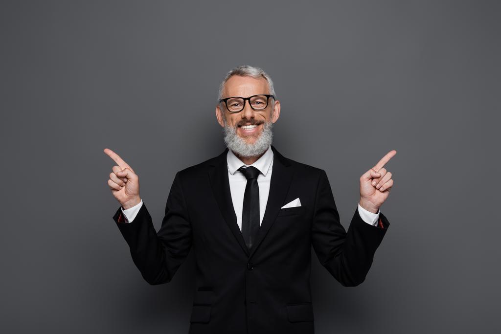 positive middle aged businessman in glasses pointing with fingers aside on grey - Photo, Image