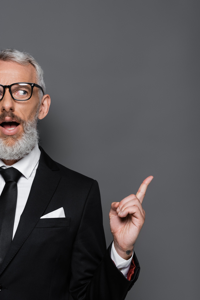 vista recortada de empresario sorprendido de mediana edad en gafas y traje apuntando con el dedo en gris - Foto, Imagen