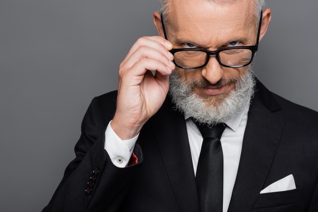 ernstige middelbare leeftijd zakenman aanpassen bril terwijl kijken naar camera geïsoleerd op grijs - Foto, afbeelding