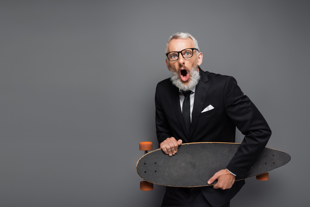 σοκαρισμένος μεσήλικας επιχειρηματίας με κοστούμι και γυαλιά κρατώντας longboard απομονωμένο σε γκρι  - Φωτογραφία, εικόνα